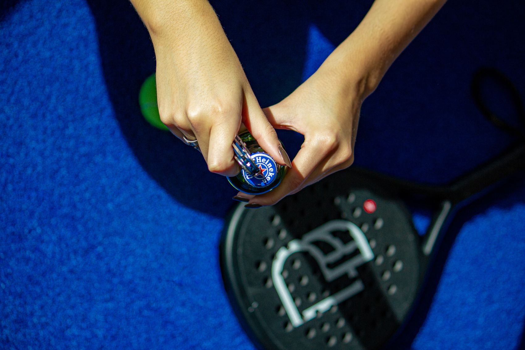 Manos abriendo una botella sobre una pista de pádel azul